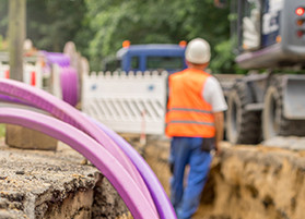 Im Vorergrund ragen fliederfarbene Gasfaserkabel in eine Baustelle, im HIntergrund steht ein Bauarbeiter mit dem Rücken zu Betrachtenden in der Baugrube.