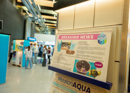 Im Vordergrund eine zeitungsähnliche Publikation der Ausstellung, im Hintergrund sind unscharf junge Ausstellungsbesucher zu sehen