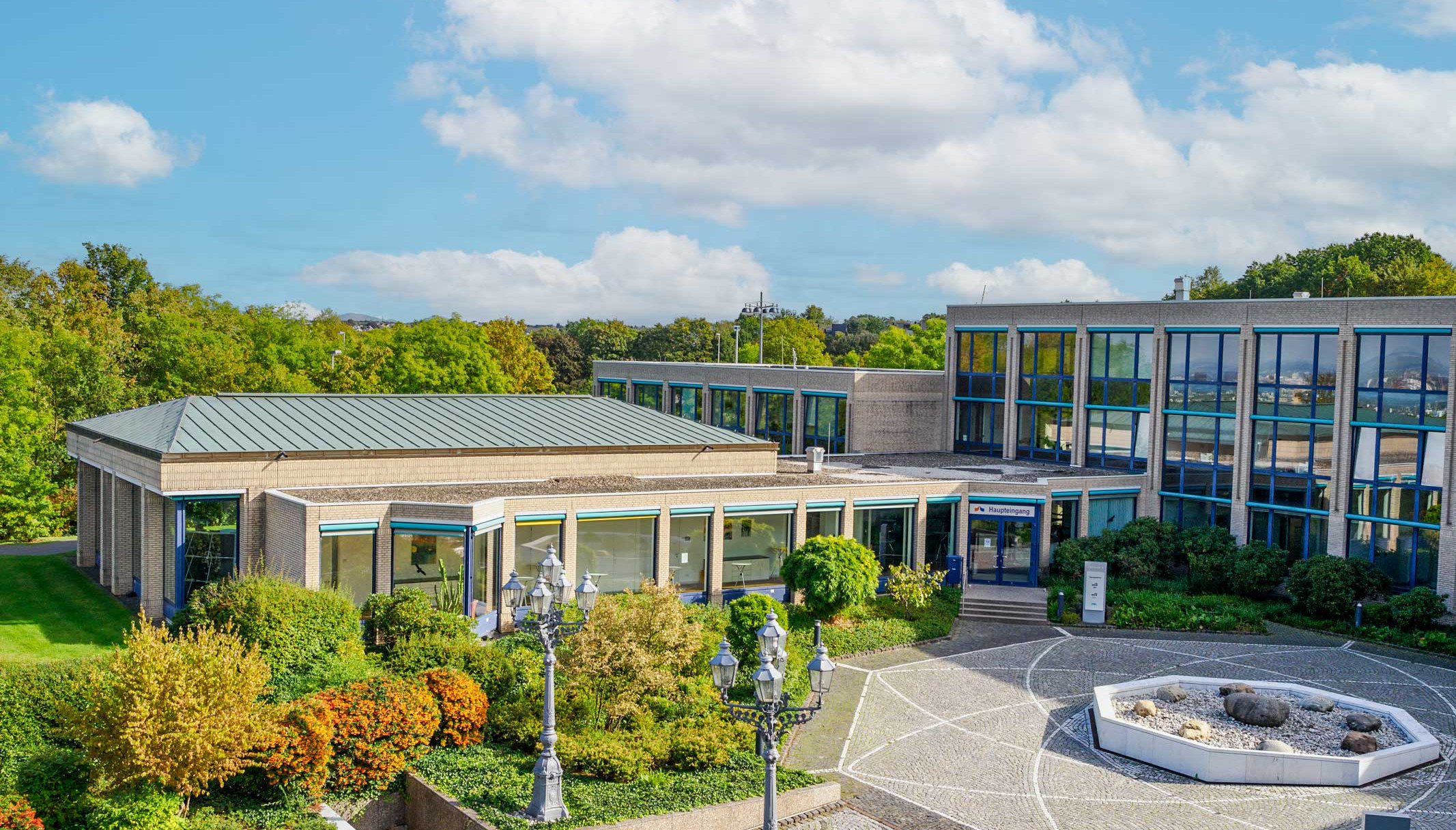 Blick von einem erhöhten Standpunkt auf das Tagungszentrum des DVGW, davor ein kleiner gepflasterter Platz mit einem Springbrunnen, rings herum viel Grün