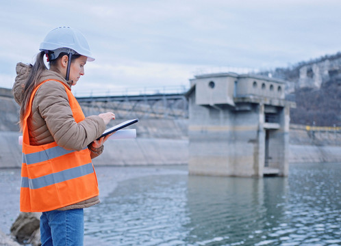 Ingenieurin mit Helm und Warnweste steht an Talsperre und prüft Daten auf ihrem Tablet