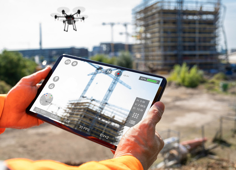 Männernhände halten ein Tablet, mittels dessen eine Drohe gesteuert wird. Im Hintergrund ist ein eingerüstetes Hochhaus zu sehen.