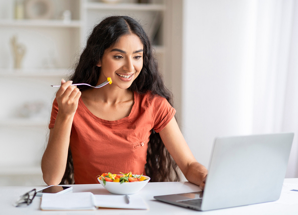 Junge Frau sitzt vor einem Laptop, während sie einen Salat isst.