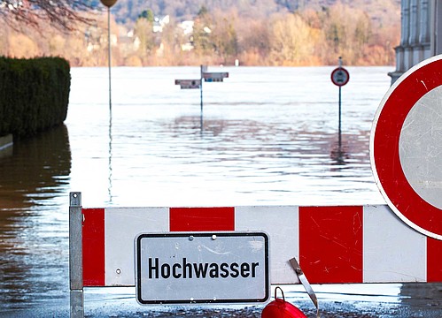 Überflutete Landschaft mit aus dem Wasser ragenden Verkehrsschildern und einer Sperre mit der Aufschrift "Hochwasser"