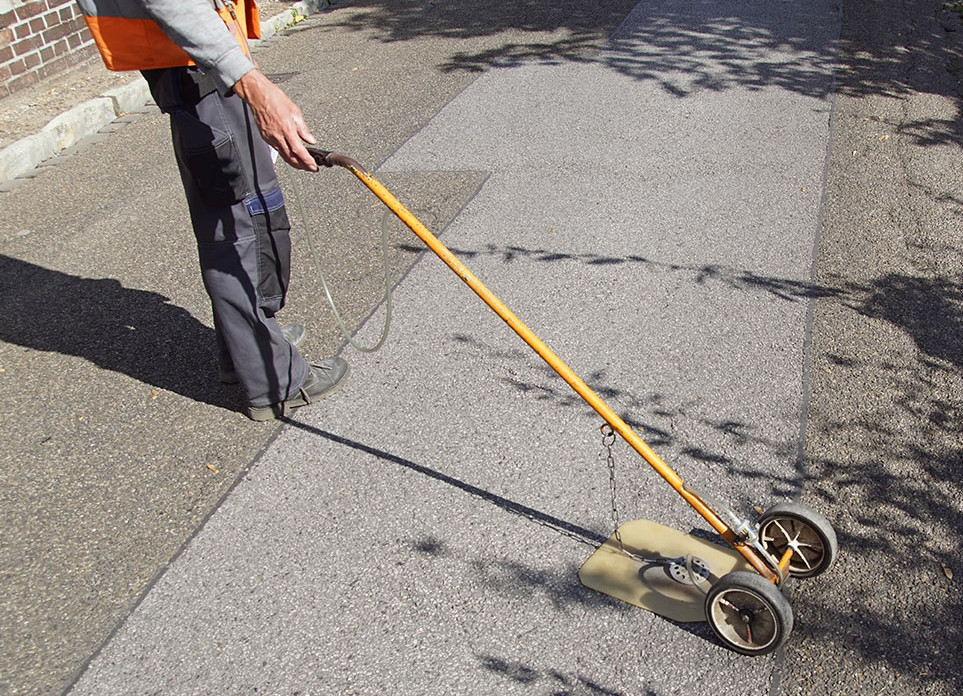 Arbeiter sucht mit einem Gerät den Boden nach Gaslecks ab. Das Gerät besteht aus einer langen Führstange mit zwei kleinen Rädern am ende, von dem an einer Kette eine flache Platte waagerecht über dem Boden schwebt.