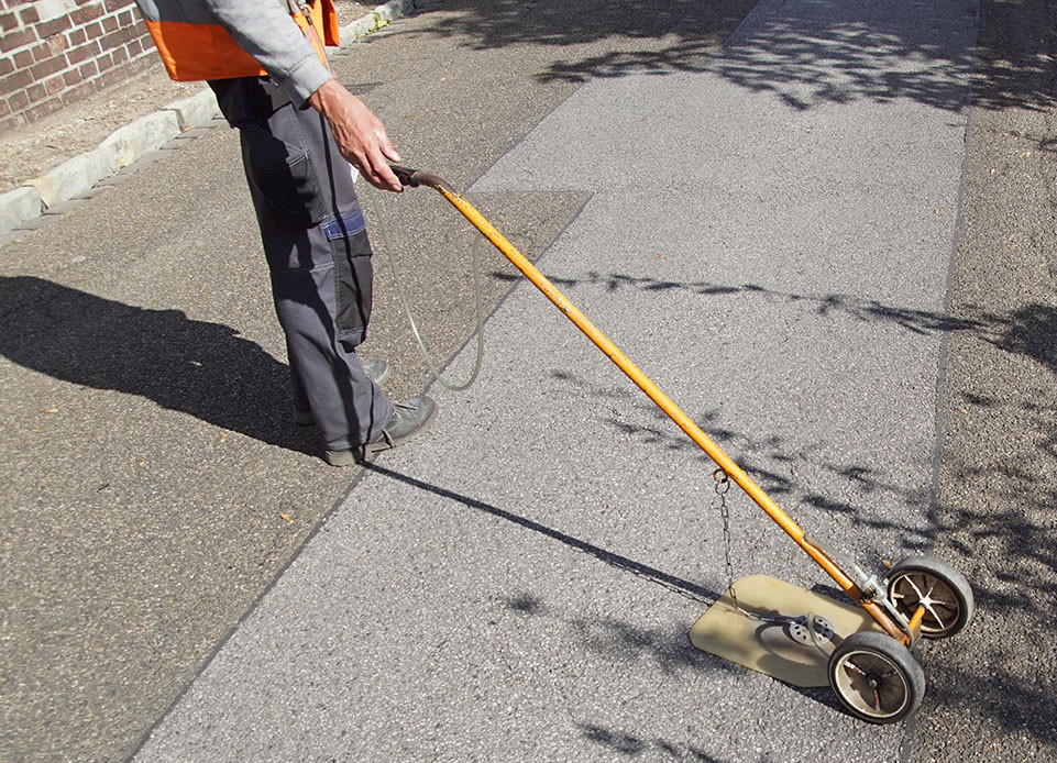 Arbeiter sucht mit einem Gerät den Boden nach Gaslecks ab. Das Gerät besteht aus einer langen Führstange mit zwei kleinen Rädern am ende, von dem an einer Kette eine flache Platte waagerecht über dem Boden schwebt.