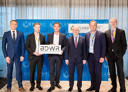 Sechs Herren in Anzügen stehen nebeneinander, zwei halten ein Schild mit der Aufschrift "BDWR Bund der Wasserstoffregionen"