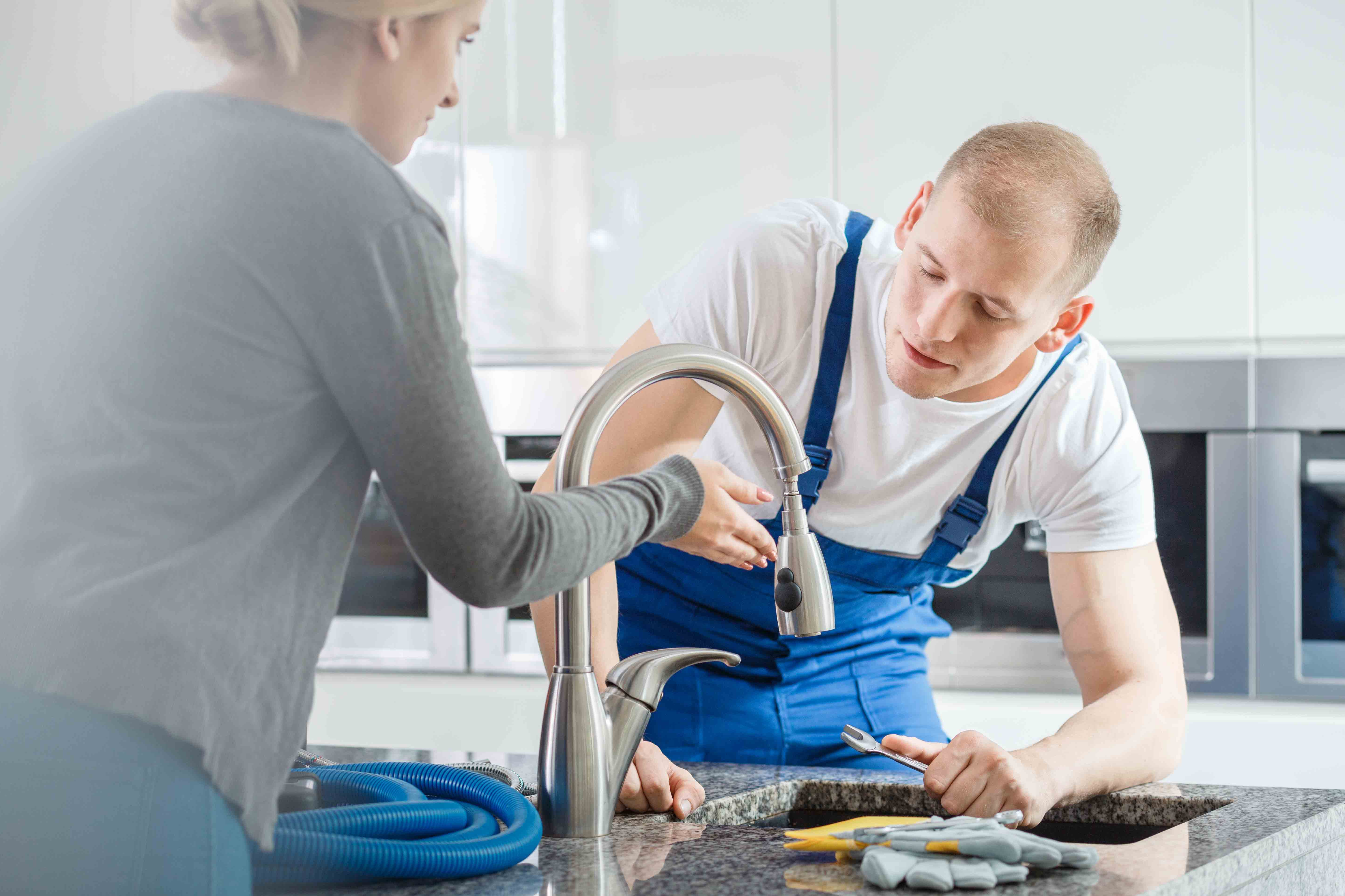 Ein Installateur arbeitet an einer Küchenarmatur, eine Frau zeigt ihm einen Fehler