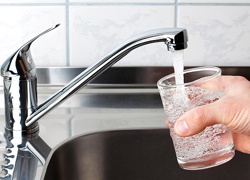 Eine Hand mit einem Wasserglas rechts im Bild, links ein geöffneter Wasserhahn, aus dem Leitungswasser ins Glas fließt.