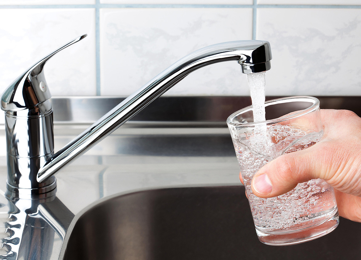 Eine Hand mit einem Wasserglas rechts im Bild, links ein geöffneter Wasserhahn, aus dem Leitungswasser ins Glas fließt.