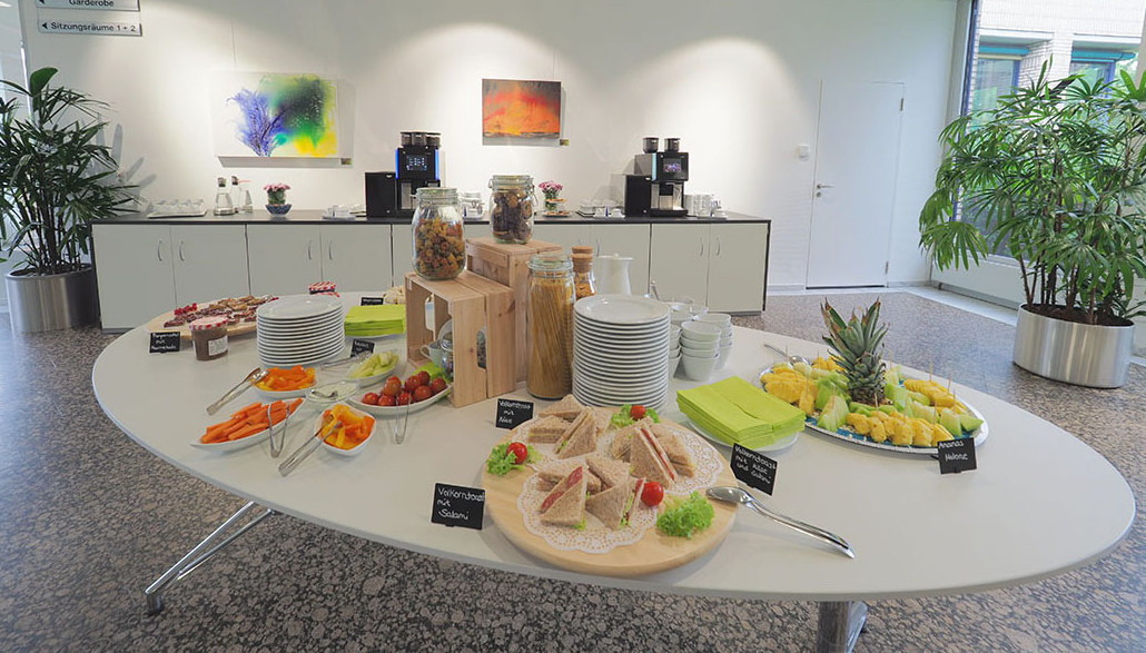 Im Vordergrund ein ovaler weißer Tisch mit Obst, Gemüse, Dips und Schnittchen, im Hintergrund ein langgezogenes Sideboard mit zwei großen Kaffeeautomaten