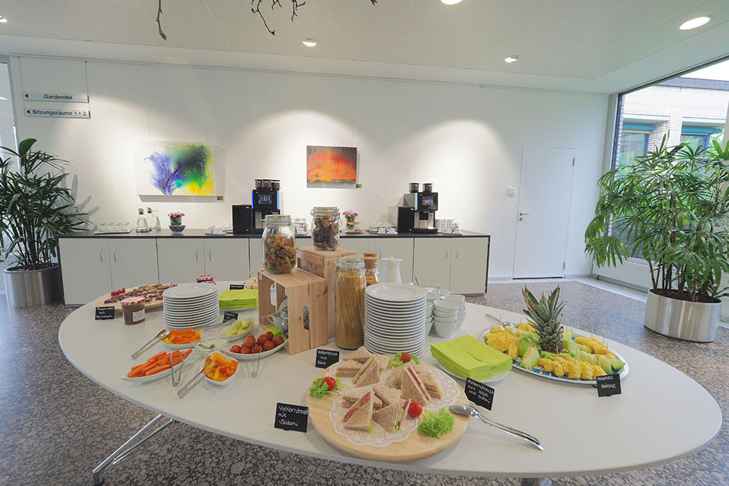 Im Vordergrund ein ovaler weißer Tisch mit Obst, Gemüse, Dips und Schnittchen, im Hintergrund ein langgezogenes Sideboard mit zwei großen Kaffeeautomaten